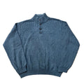 A dusty slate blue knit mock-neck sweater with three dark buttons, a white background. A pair of bare feet is visible at the bottom edge of the frame.