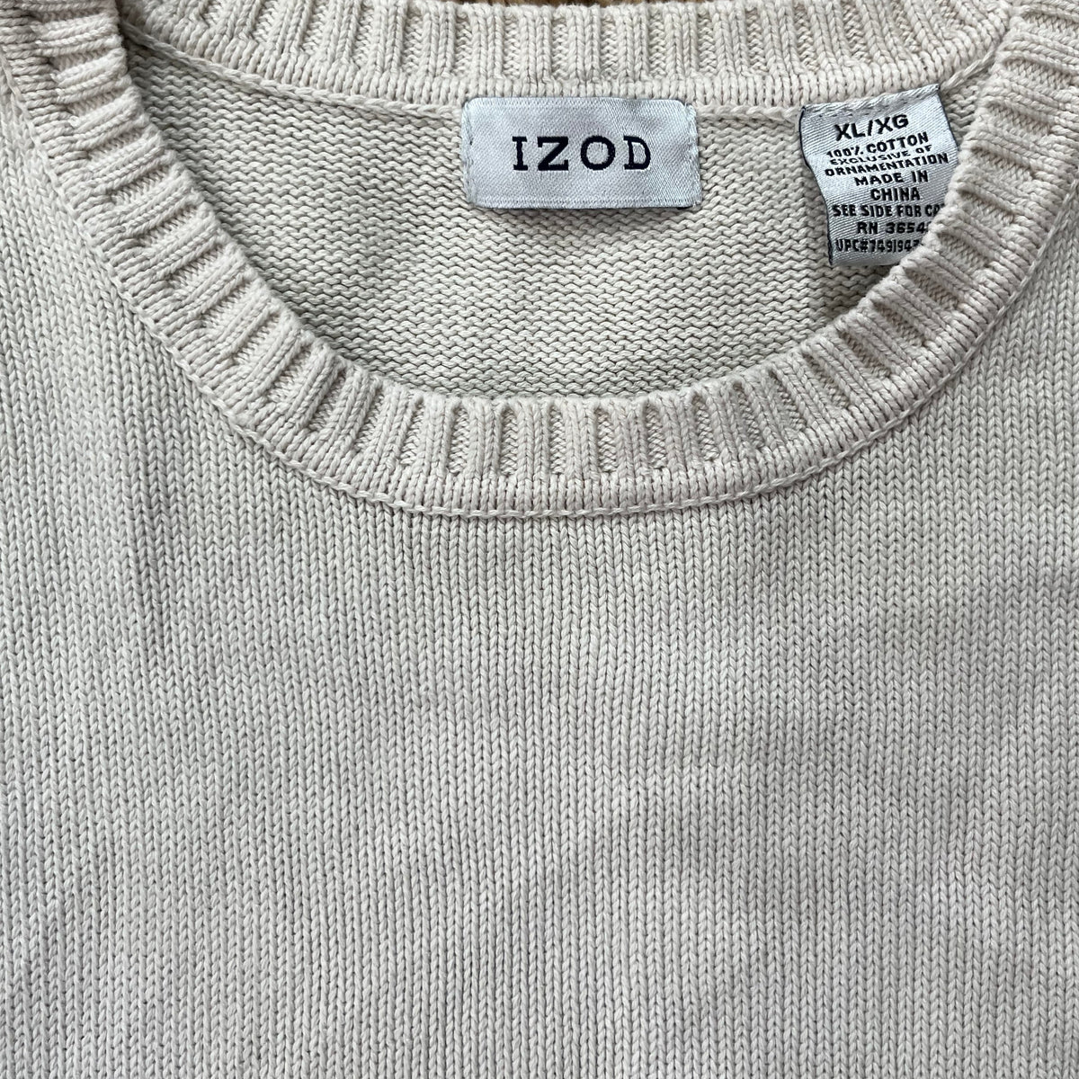 Close-up of a light beige knit crewneck sweater with a ribbed collar, featuring an 'IZOD' brand label and a size tag, laid flat on a brown shag rug.