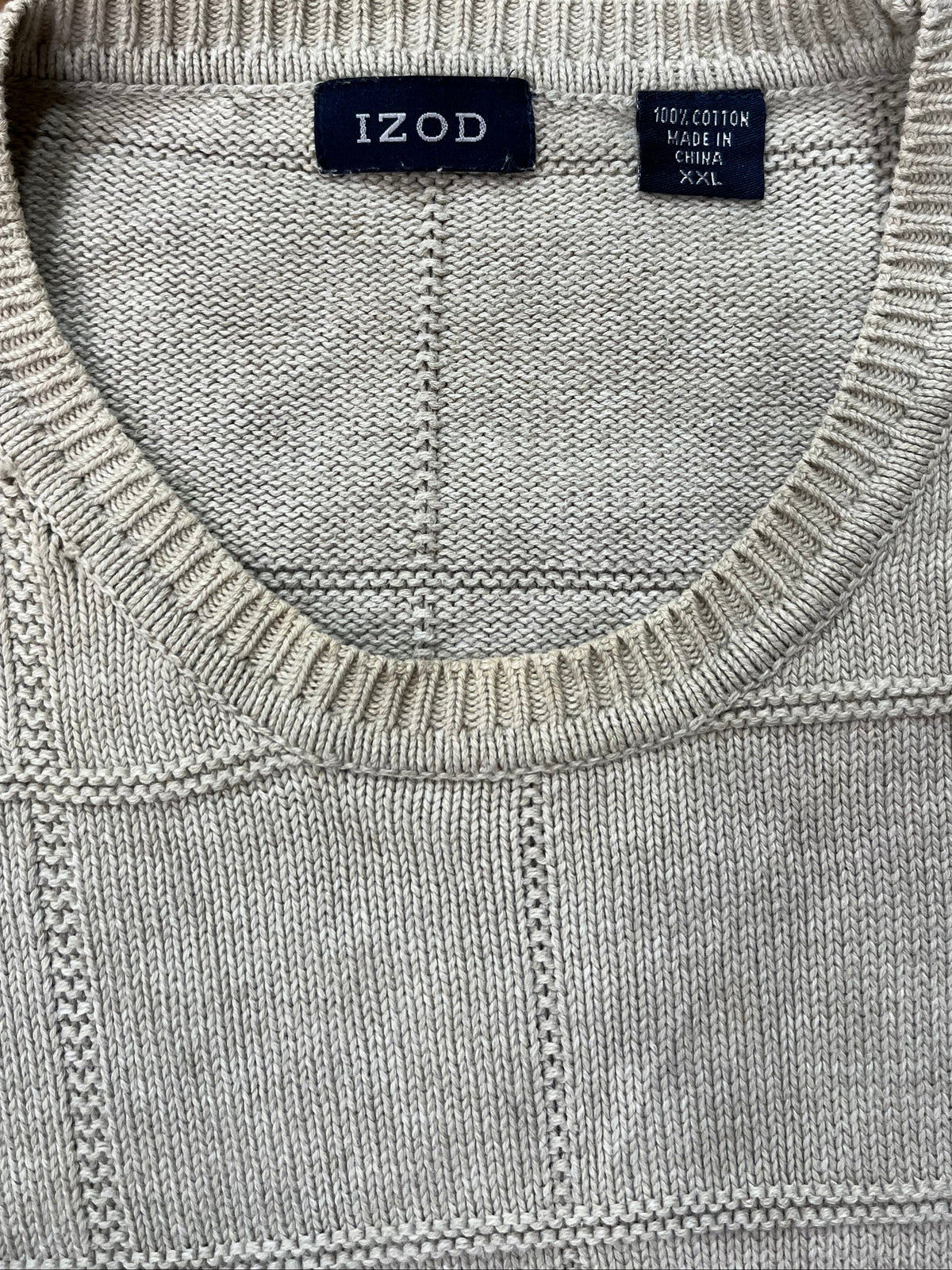 A close-up of a light gray knit sweater with a crew neck, laid flat on a beige shag rug. The brand label 'IZOD' is visible inside the collar.