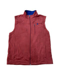 A sleeveless zip-up vest in a muted brick red color, laid flat on a beige shag rug. The vest features a blue inner collar lining and a small blue logo on the left chest pocket.
