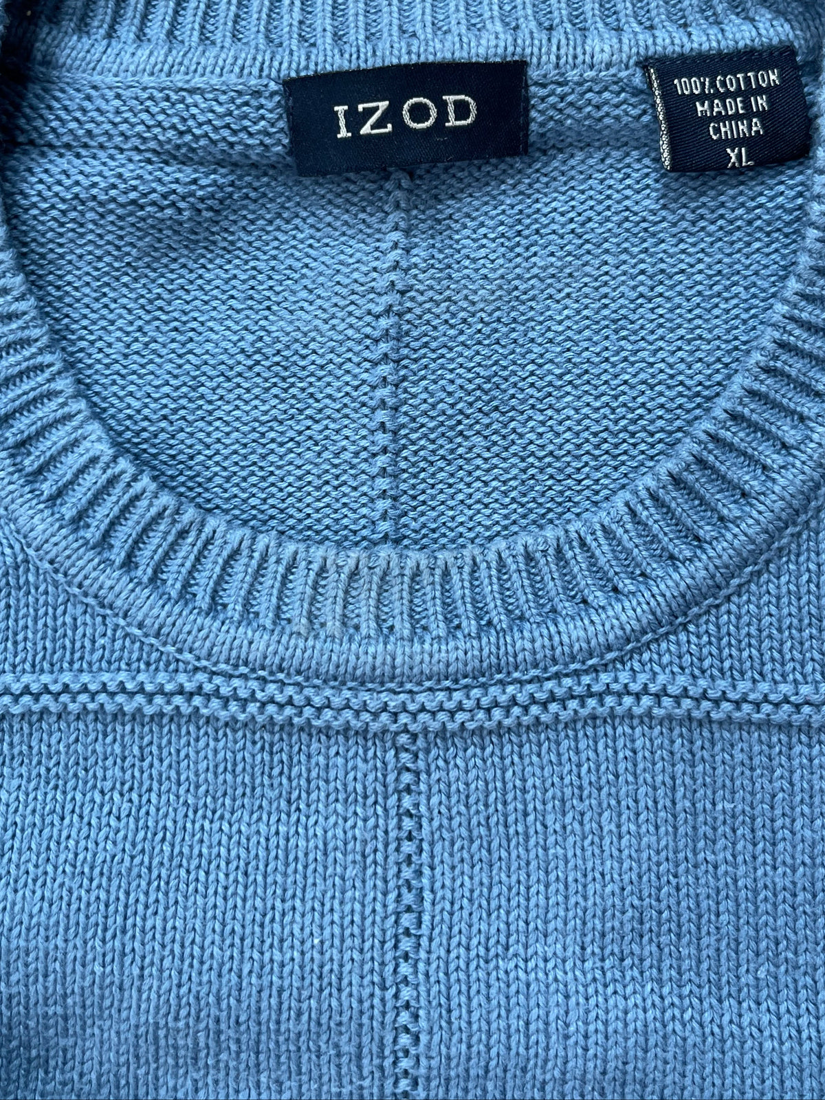 Close-up of a light blue knit sweater showing the ribbed crew neck with an 'IZOD' brand label and a '100% COTTON MADE IN CHINA YI' tag, resting on a beige shag rug.