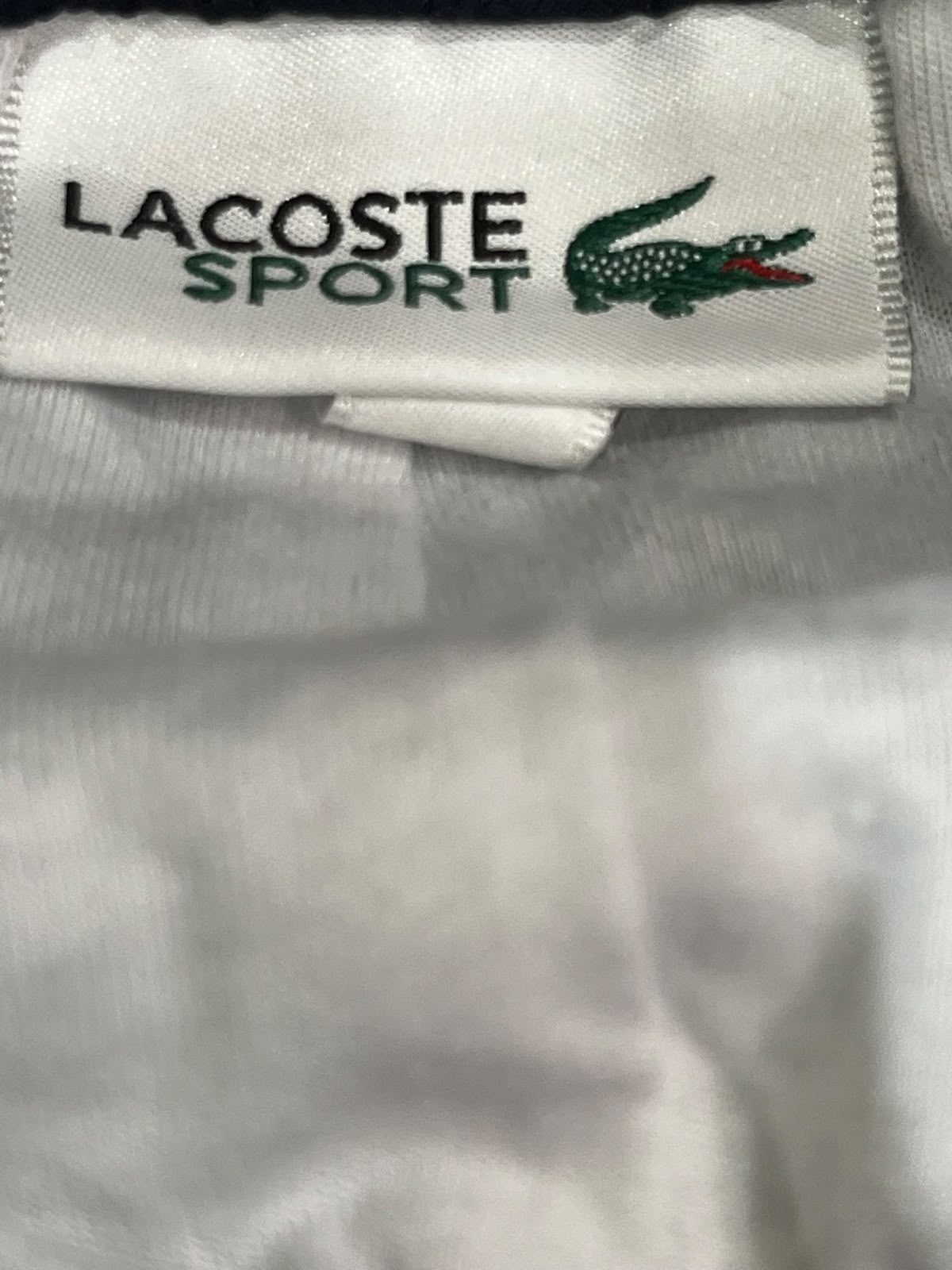 Close-up of a Lacoste Sport garment label sewn onto white fabric, showing the black and green text logo with the crocodile emblem.