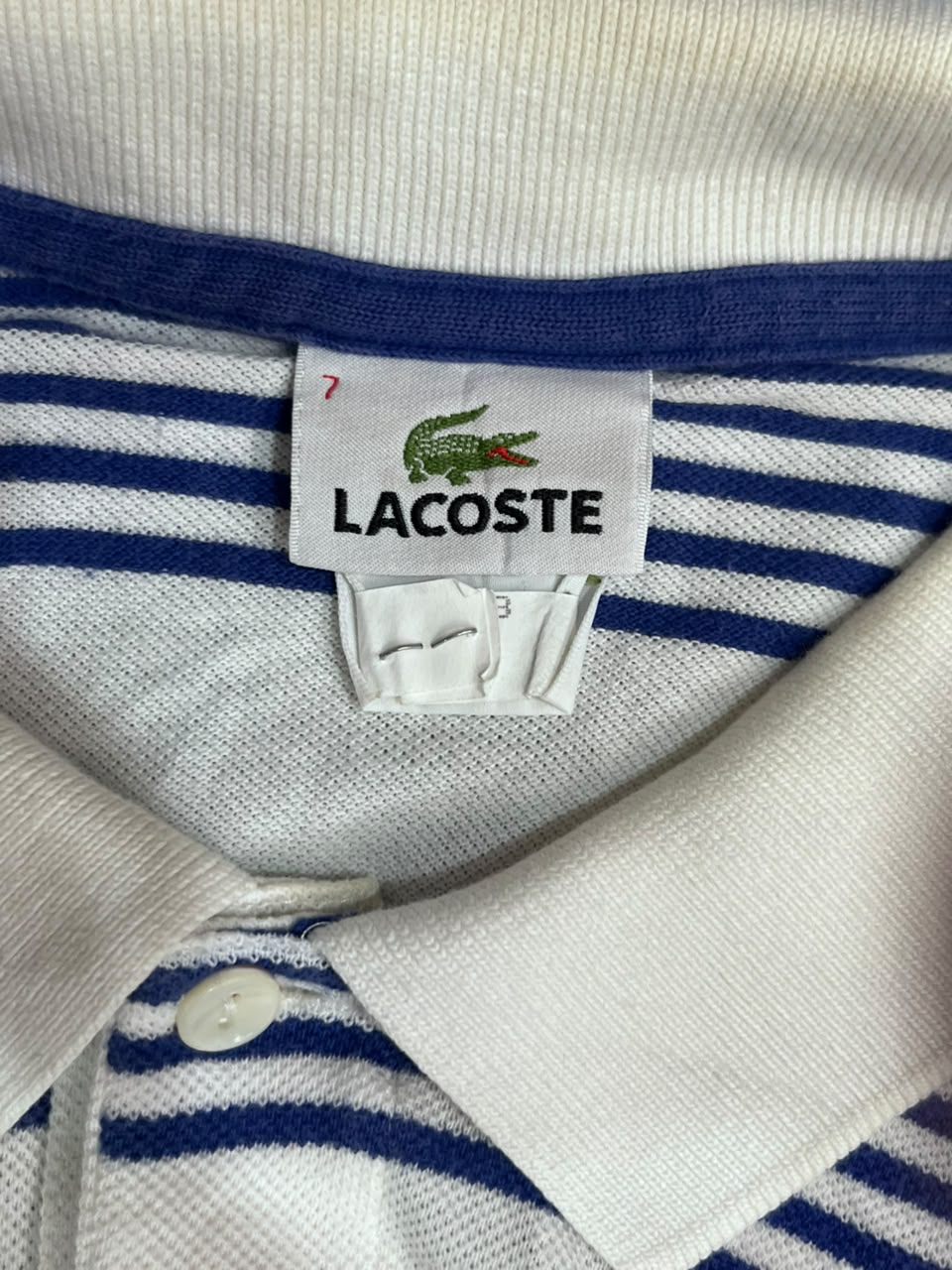 Close-up of a Lacoste polo shirt collar and neck label, showing a white ribbed knit collar, navy blue and white horizontal stripes, and the Lacoste crocodile logo on a white fabric tag.