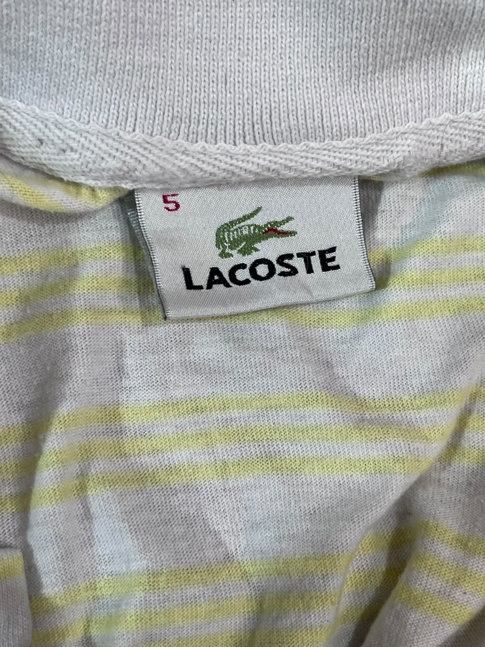 Close-up of a Lacoste garment neck label on a white and pale yellow striped knit top, with the green crocodile logo and the number 5 visible.