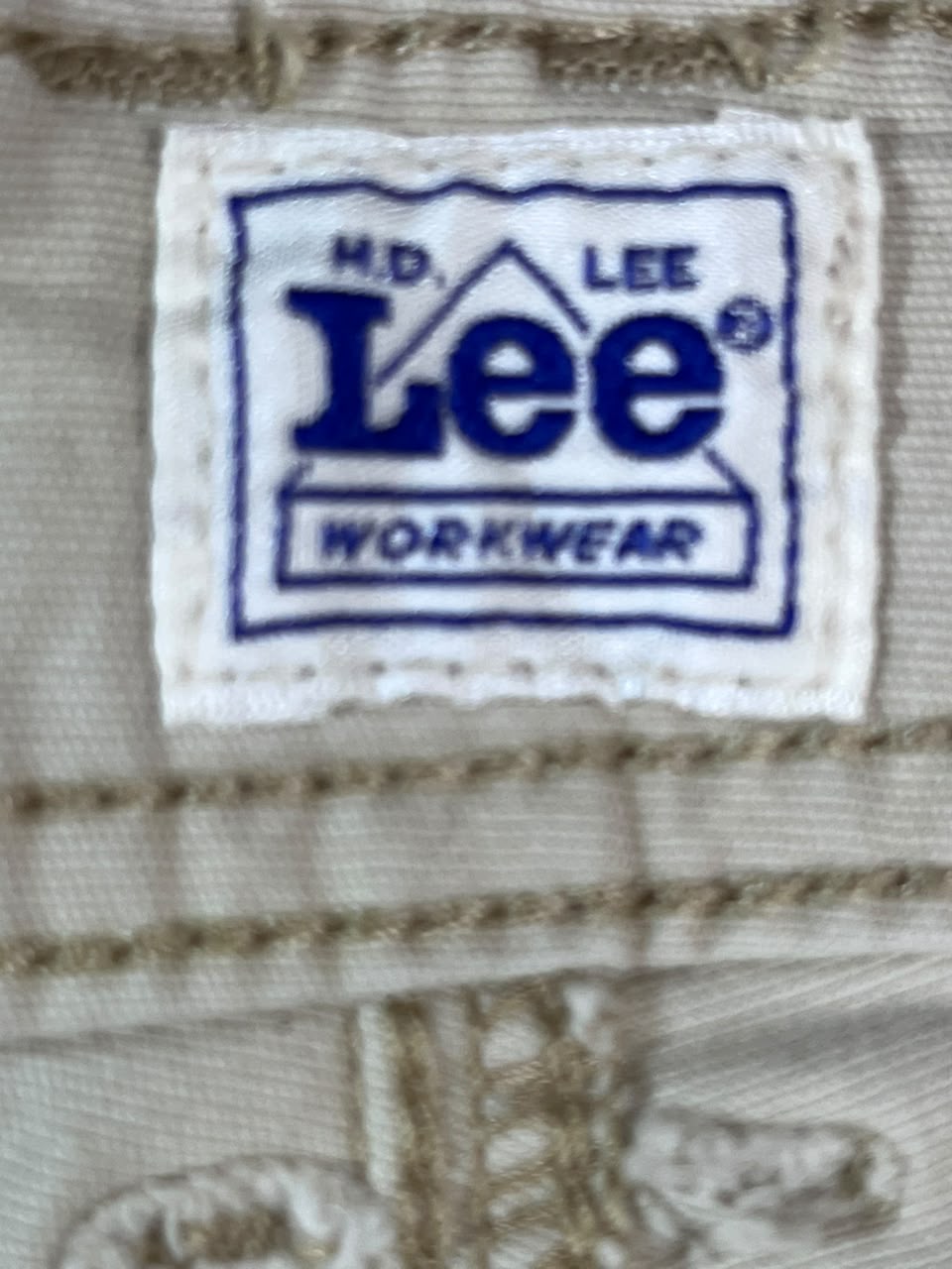 Close-up of a beige woven fabric label patch with blue text reading 'M.D. LEE Lee WORKWEAR' stitched onto the material.