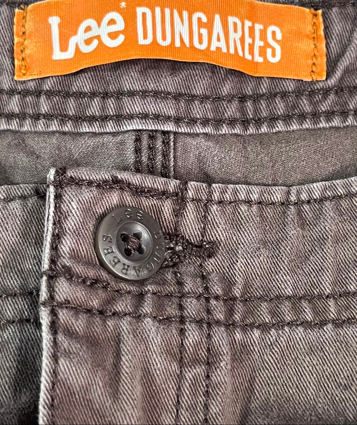 Close-up of Lee Dungarees jeans showing the orange brand label at the waistband and a gray denim button on the pocket.