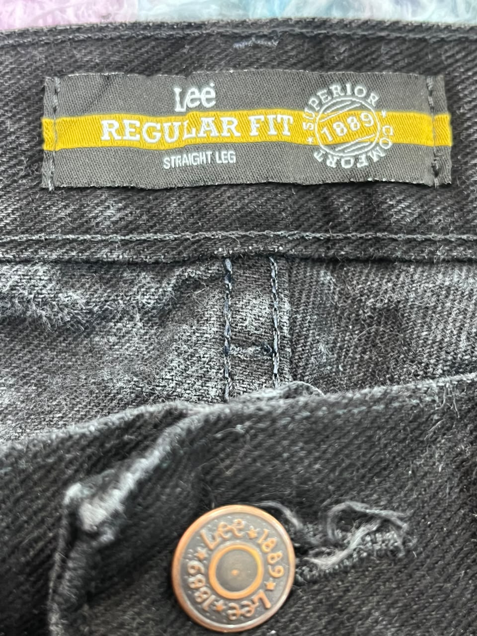 Close-up of the waistband of Lee Regular Fit straight-leg jeans in a washed black or charcoal denim, featuring a yellow and gray brand label and a bronze button.