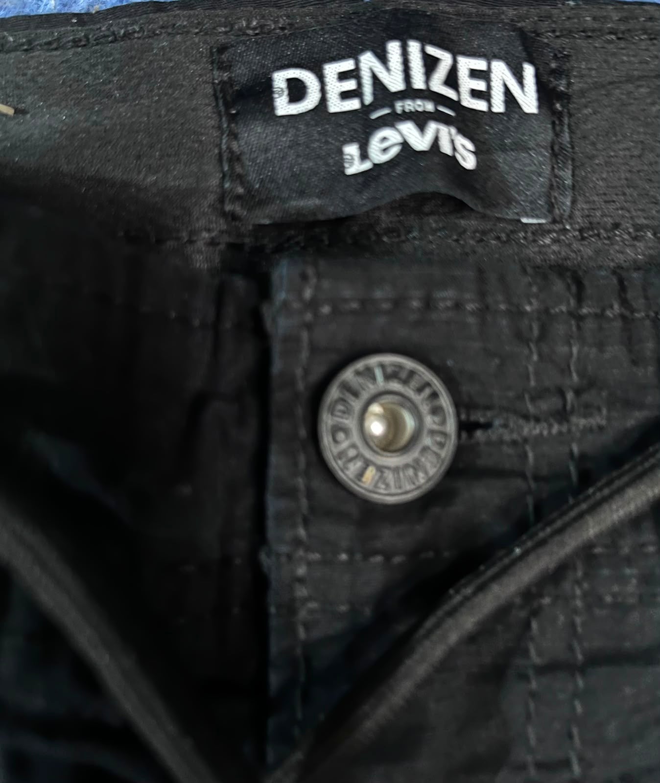 Close-up of black denim pants showing the interior waistband label that reads 'DENIZEN from Levi's' and a metal button on the pocket.