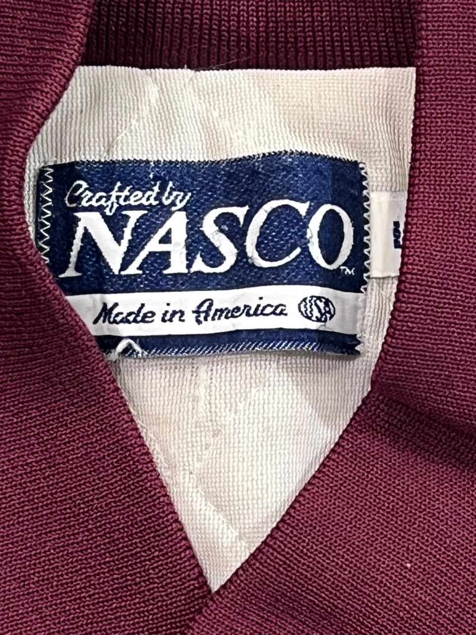 Close-up of a maroon knit garment's interior neck label reading 'Crafted by NASCO' and 'Made in America' on a white background.