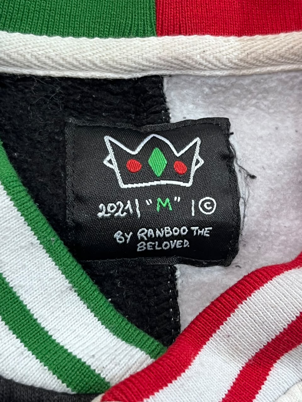 Close-up of a knit garment label reading '2021 "M" I© BY RANBOO THE BELOVER' with a crown graphic, shown inside a striped garment with green, white, black, and red sections.
