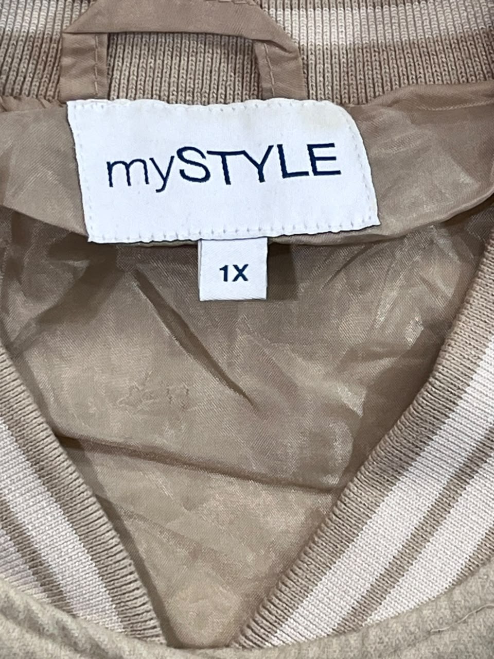 Close-up of a beige knit garment with a white inner label reading 'mySTYLE' and a smaller size tag reading '1X'.