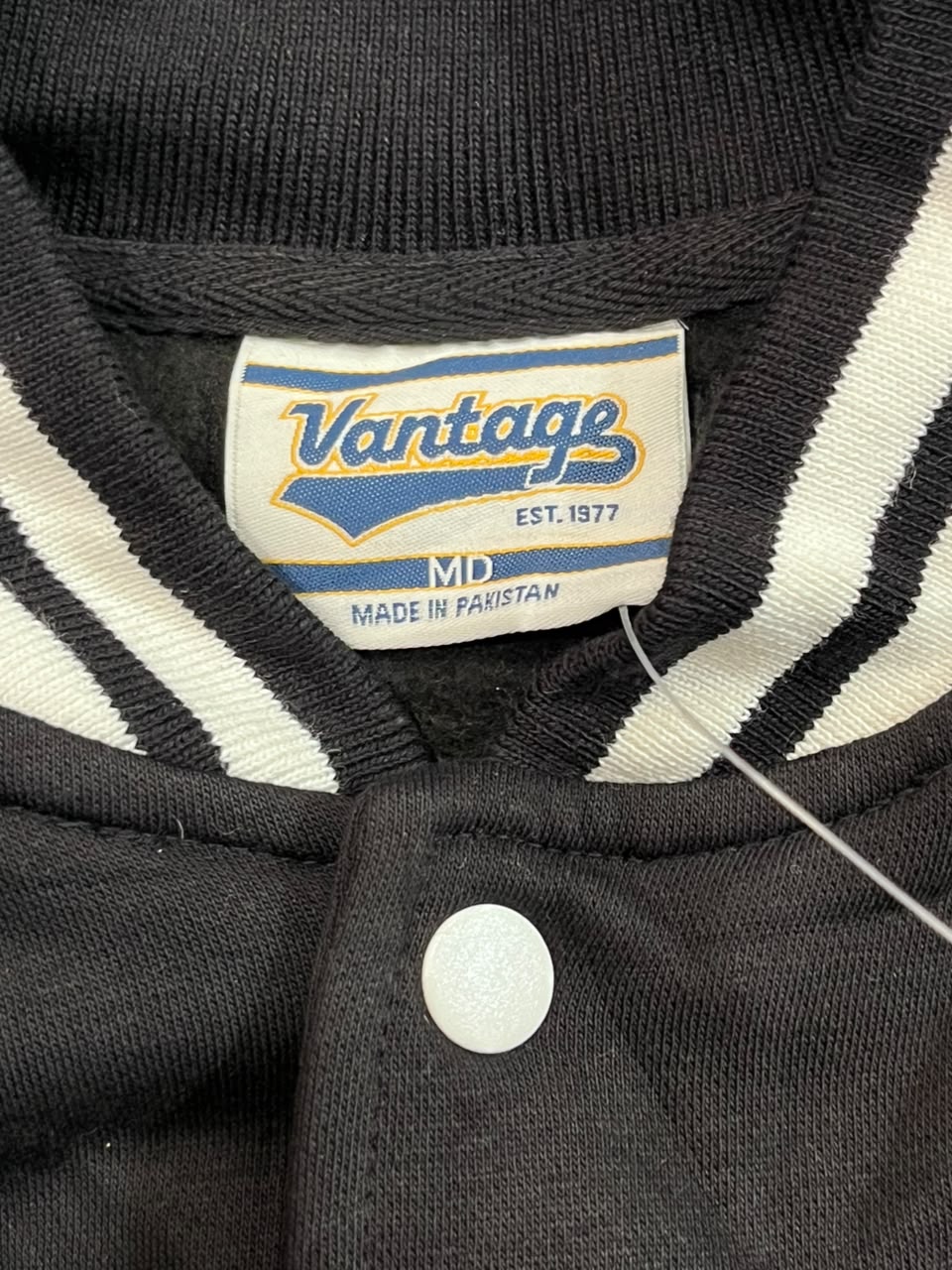 Close-up of a Vantage brand varsity-style jacket collar showing a cream label with blue and yellow branding, black and white striped ribbed knit trim, and a white snap closure.