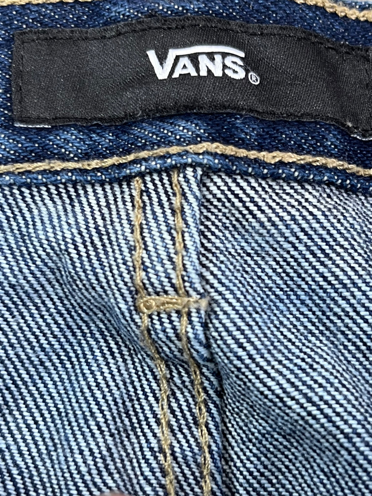 Close-up of a Vans denim garment showing the black Vans brand label stitched onto the waistband, with blue twill fabric and tan contrast stitching.