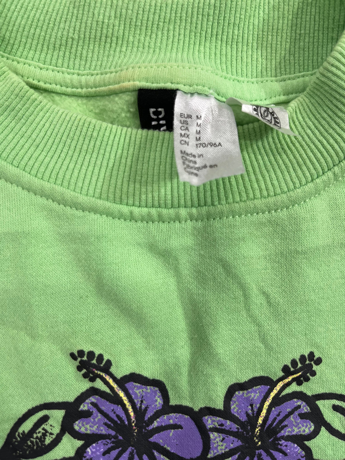 Close-up of a light green ribbed knit garment, likely a sweatshirt or sweater, featuring a purple hibiscus flower graphic on the left and a white size tag with a black brand label inside the collar.