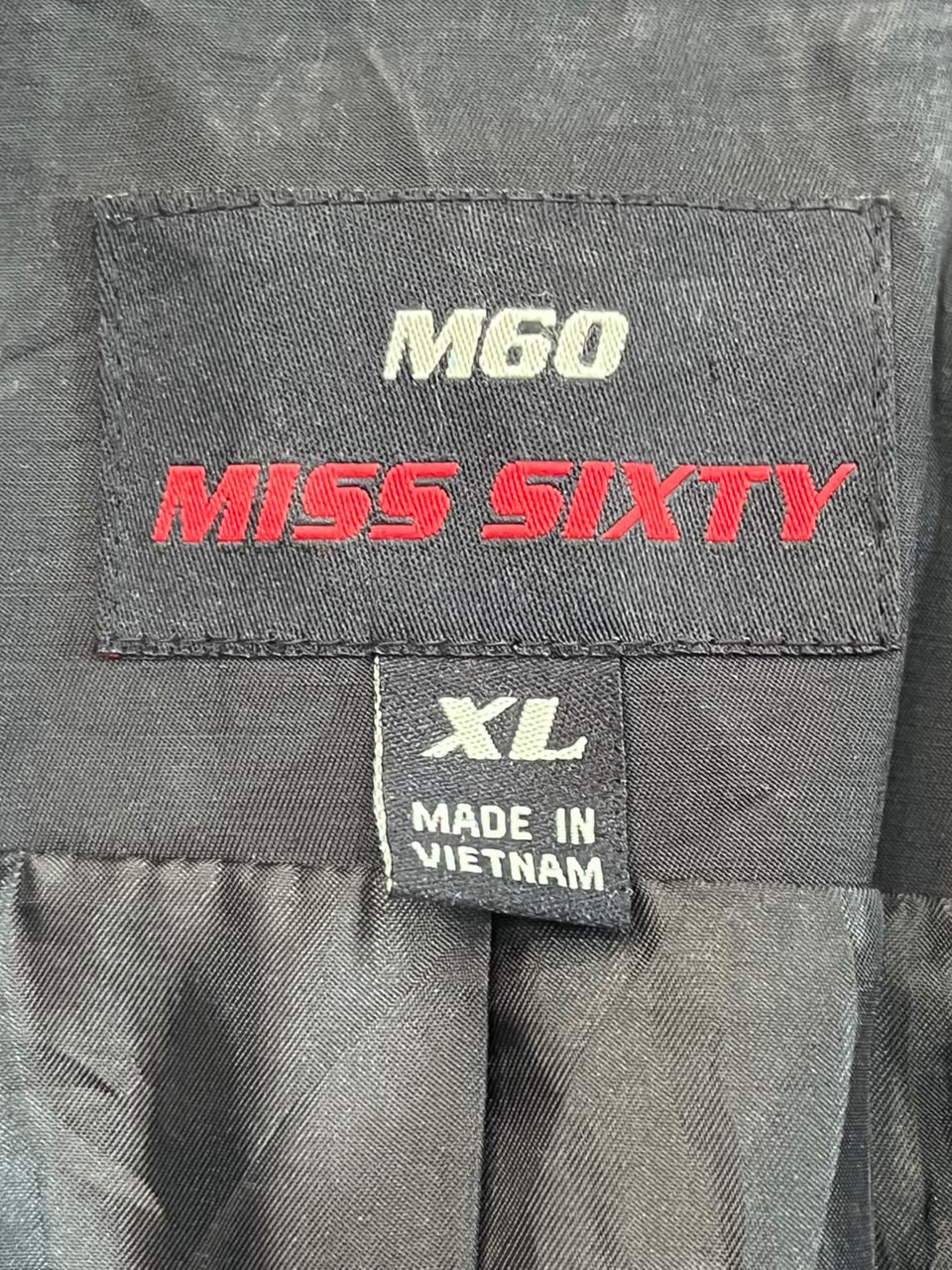 Close-up of a garment label reading M60 MISS SIXTY in white and red on a dark gray fabric, with a smaller tag below reading XL and MADE IN VIETNAM.