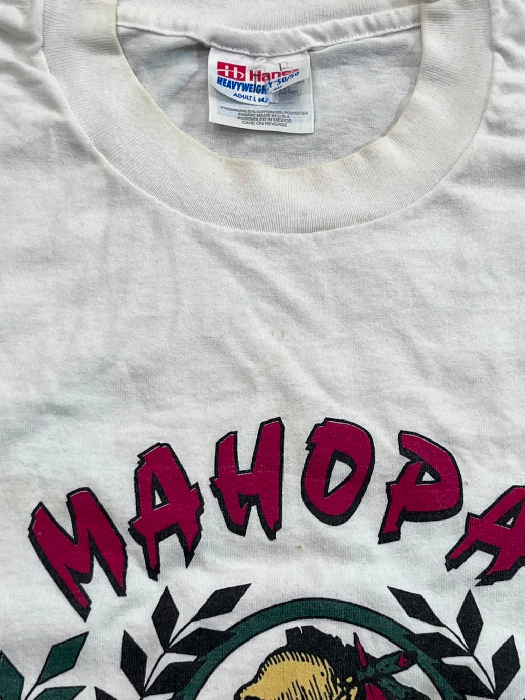 Close-up of a white cotton t-shirt featuring a vintage-style graphic with the word 'MAHOPAH' in dark red letters outlined in black, above a circular emblem with green leaves and a yellow-and-black face. The inside neck label shows 'HB Hansen' and 'CraftWash'.