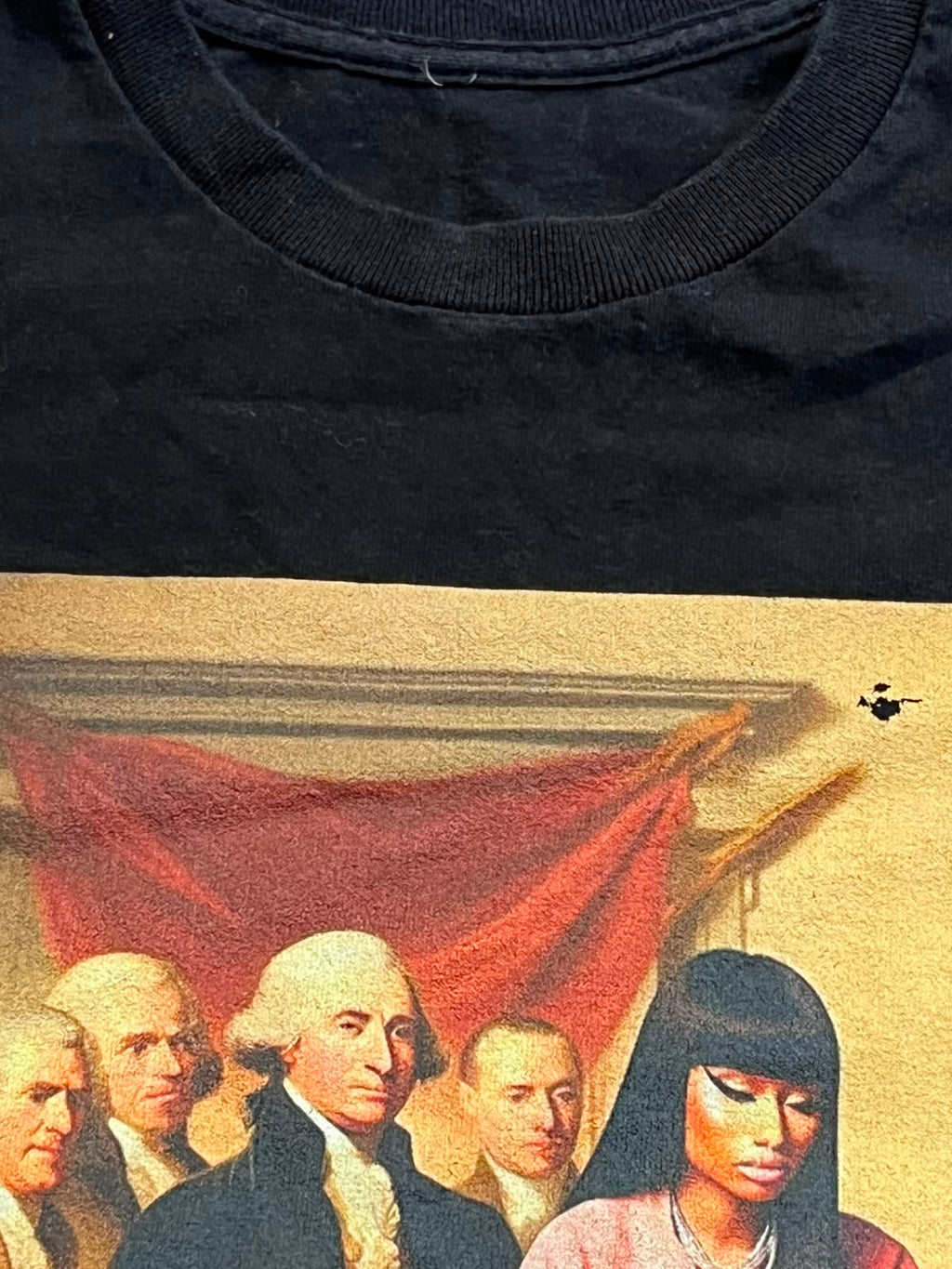 Close-up of a black crewneck t-shirt showing the collar and a portion of a vintage-style graphic print featuring a figure in a red cloak against a beige background.