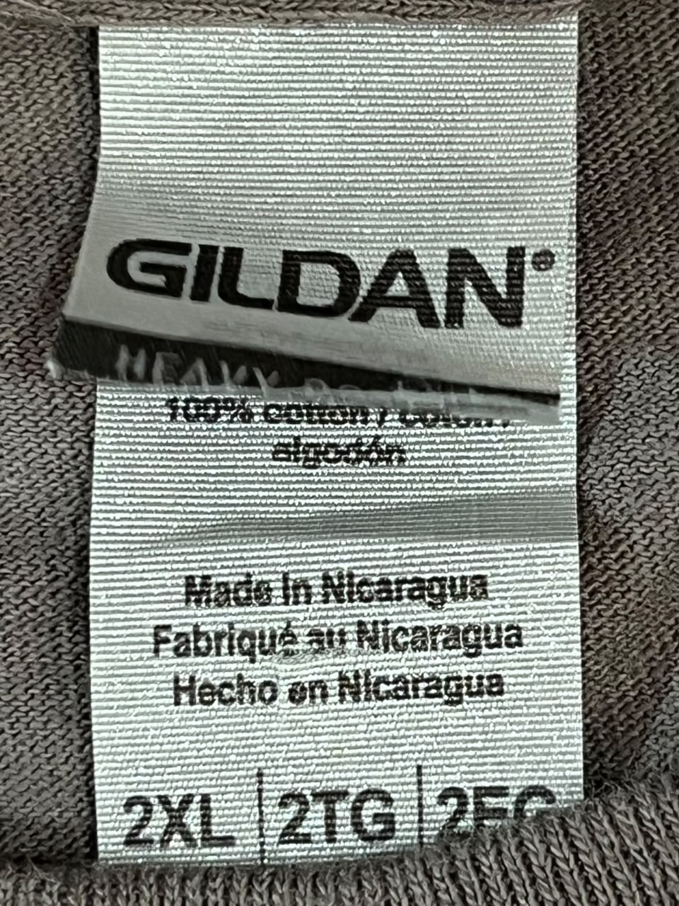 Close-up of a Gildan brand fabric care label sewn onto a gray knit garment, showing text including 'GILDAN', '100% cotton', 'Made in Nicaragua', and size information '2XL | 2TG | 2FQ'.