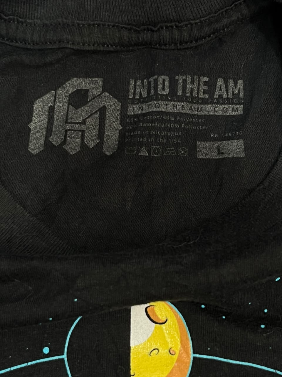 Close-up of the inside collar of a black t-shirt showing the printed neck label with 'INTO THE AM' and a stylized 'IM' logo. The bottom of the shirt features a graphic of a yellow planet with a black ring and light blue orbit lines.