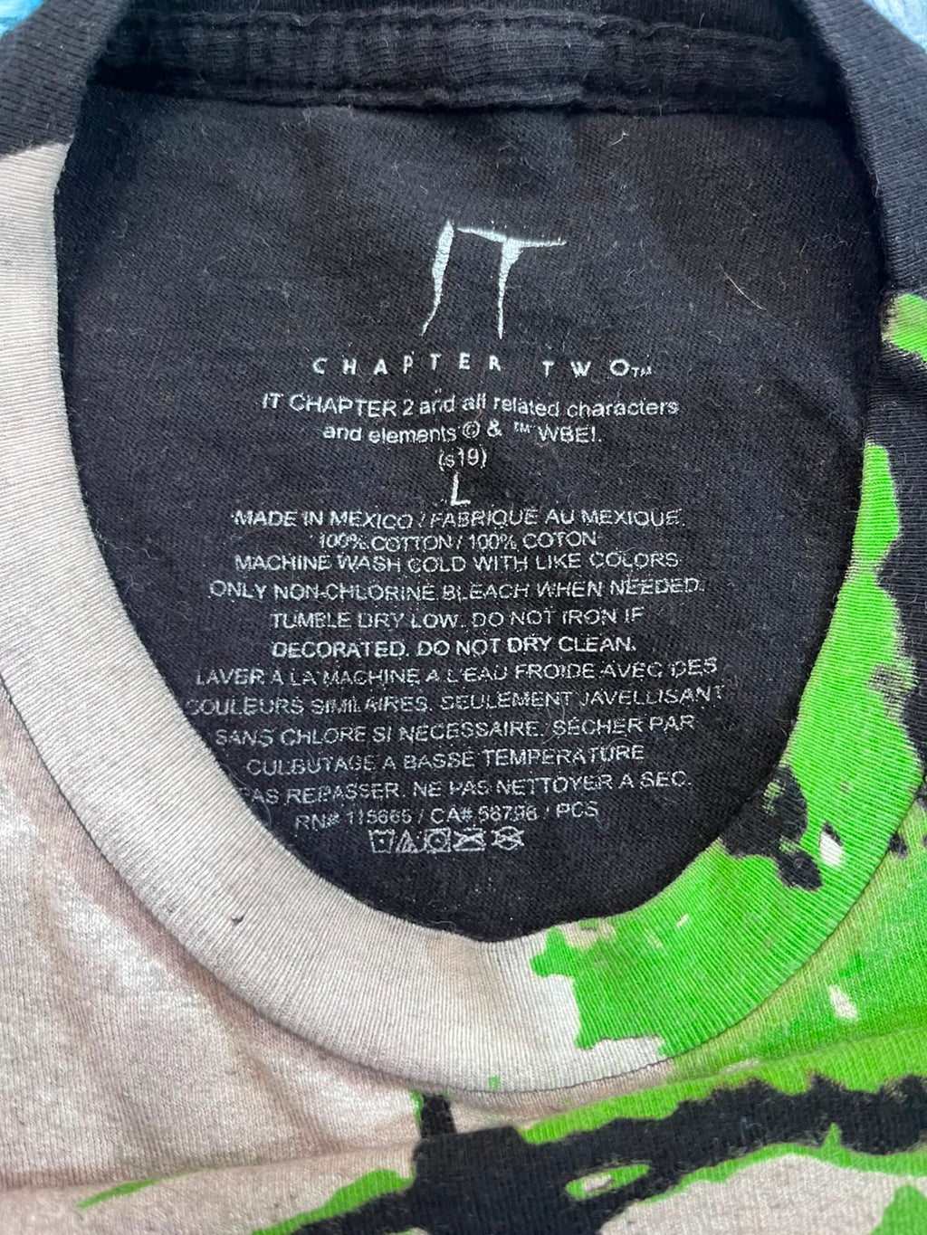 Close-up of the inside collar of a t-shirt showing printed care instructions and the brand name 'IT CHAPTER 2'. The collar is black with white text, and the surrounding shirt fabric is a light gray heather with a bright green graphic print on the shoulder.