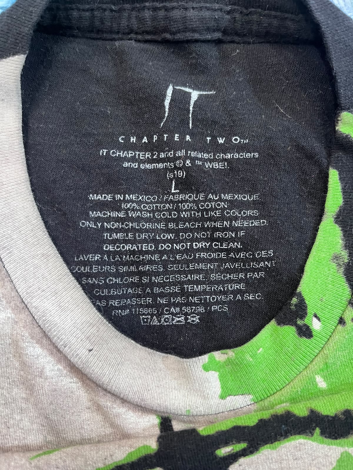 Close-up of the inside collar of a t-shirt showing printed care instructions and the brand name 'IT CHAPTER 2'. The collar is black with white text, and the surrounding shirt fabric is a light gray heather with a bright green graphic print on the shoulder.