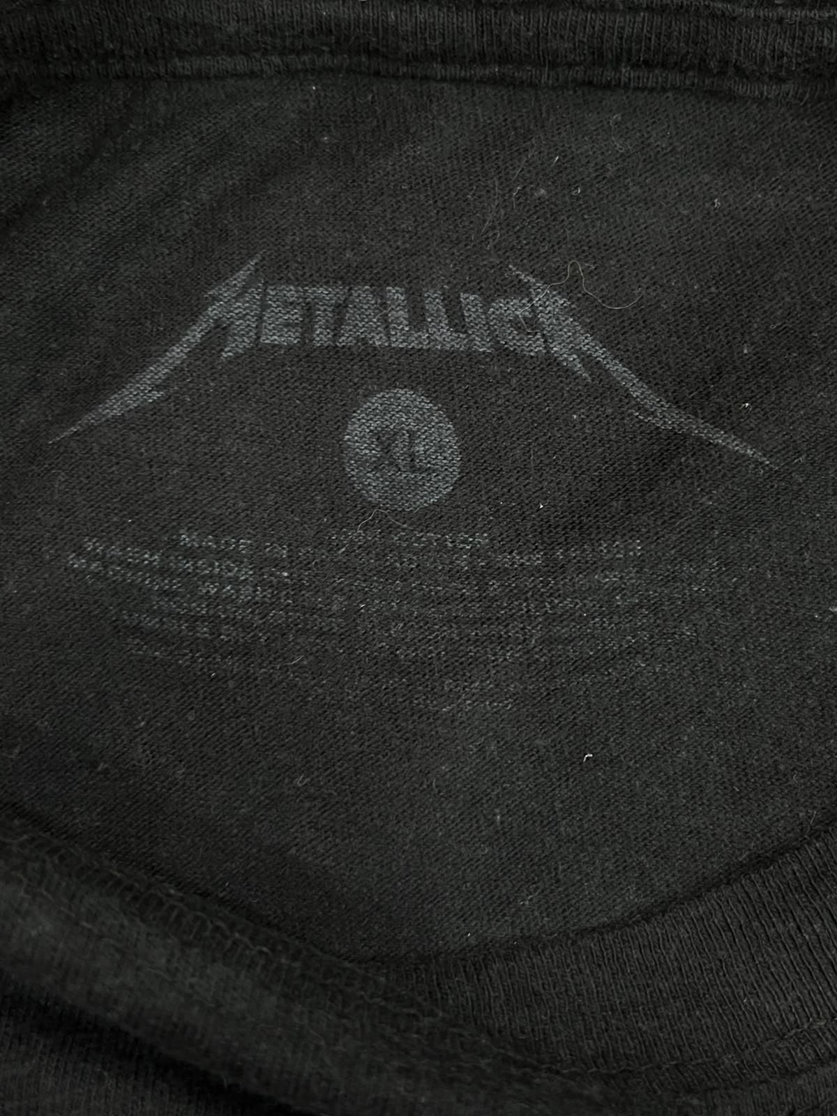 Close-up of the inside neck label of a black Metallica t-shirt, showing the band's logo and size information.