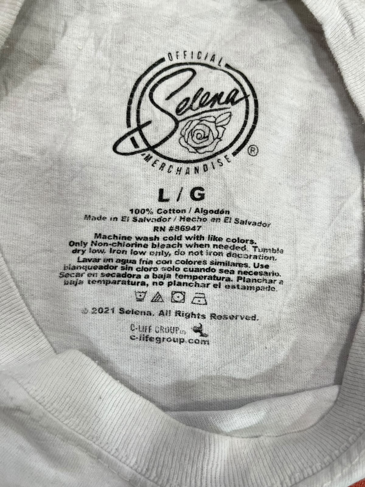 Close-up of the inside neck label of a white cotton t-shirt by Selena, showing the brand logo, 'Official Selena Merchandise' text, and care instructions.