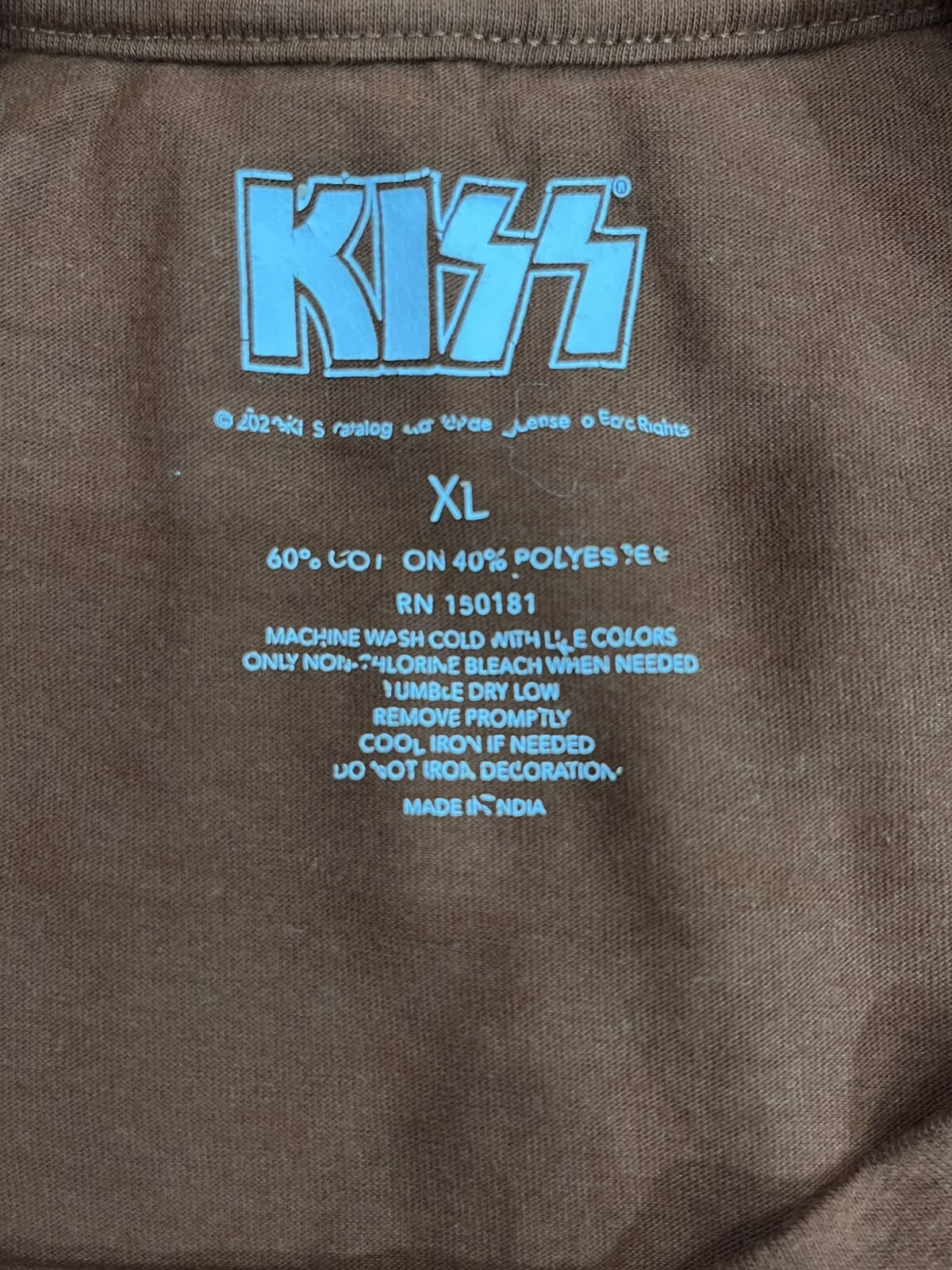 Close-up of the inside neck label of a brown garment, showing a large light blue 'KISS' logo and size information printed on the fabric.