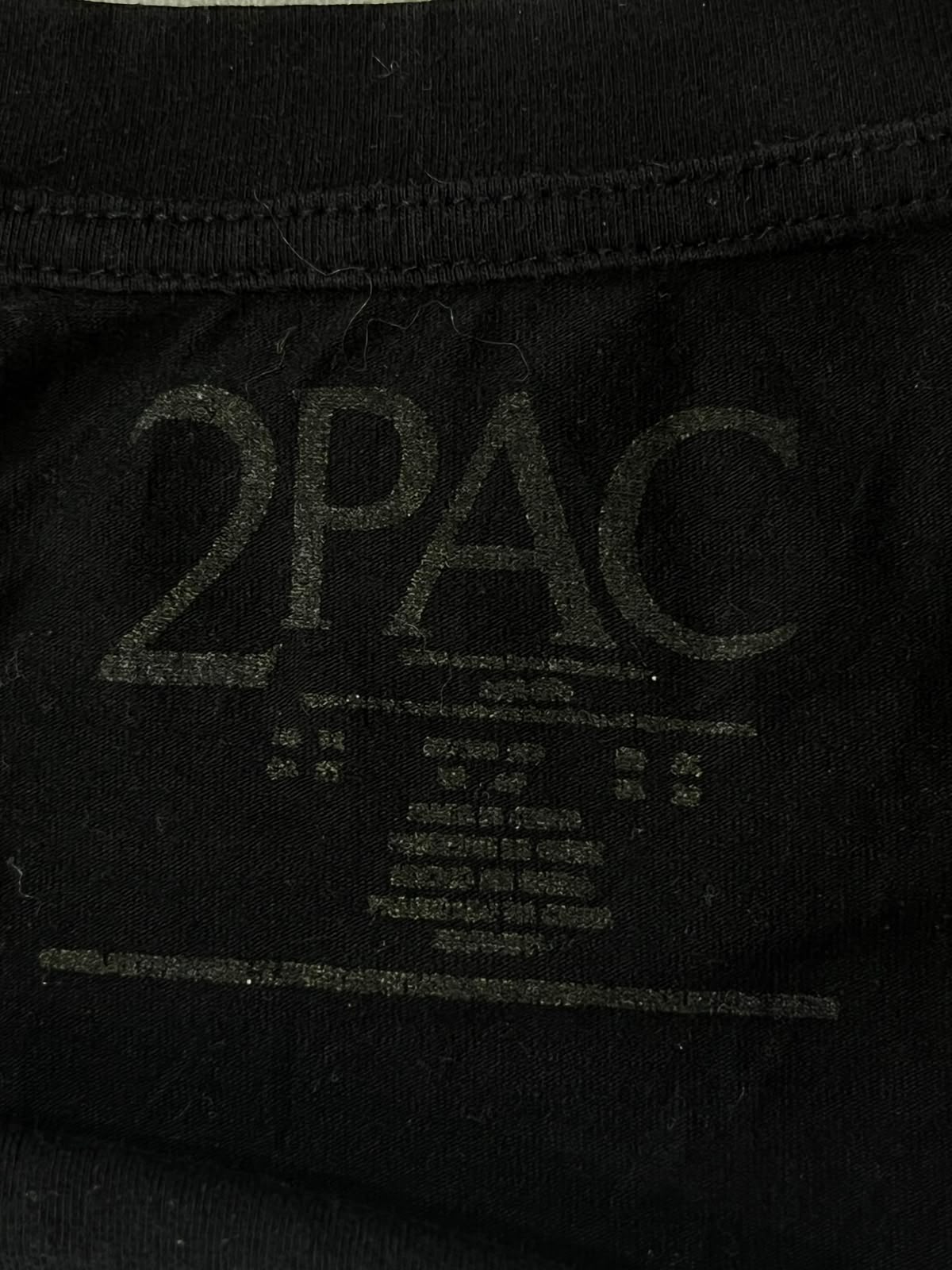 Close-up of a black garment showing a faded dark gray '2PAC' logo and small printed text inside the collar.