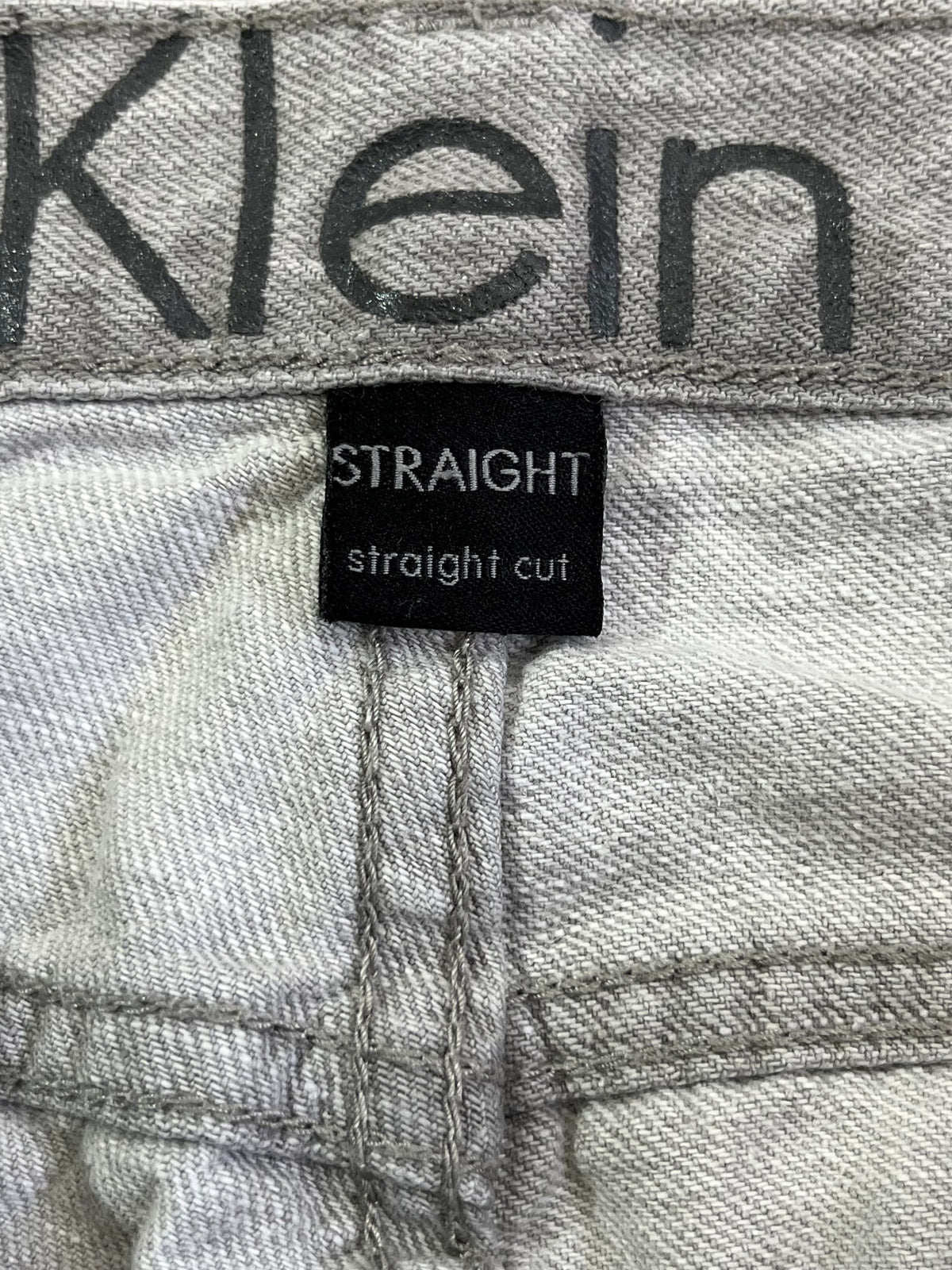 Close-up of light gray denim jeans showing the Calvin Klein waistband with the brand name printed in dark gray, and a black sewn-in label reading 'STRAIGHT straight cut'.