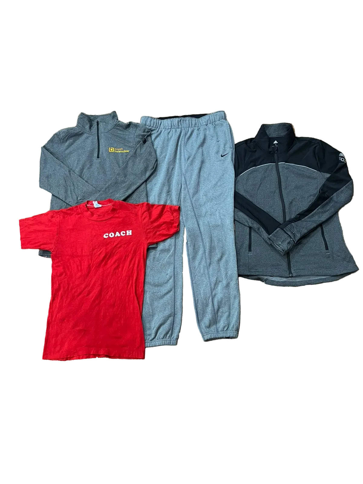 A flat lay of four clothing items on a beige shag rug: a gray Nike zip-up hoodie, gray sweatpants with a visible Nike swoosh, a red short-sleeve Coach t-shirt, and a dark gray and black quarter-zip jacket. Bare feet are visible at the bottom edge.
