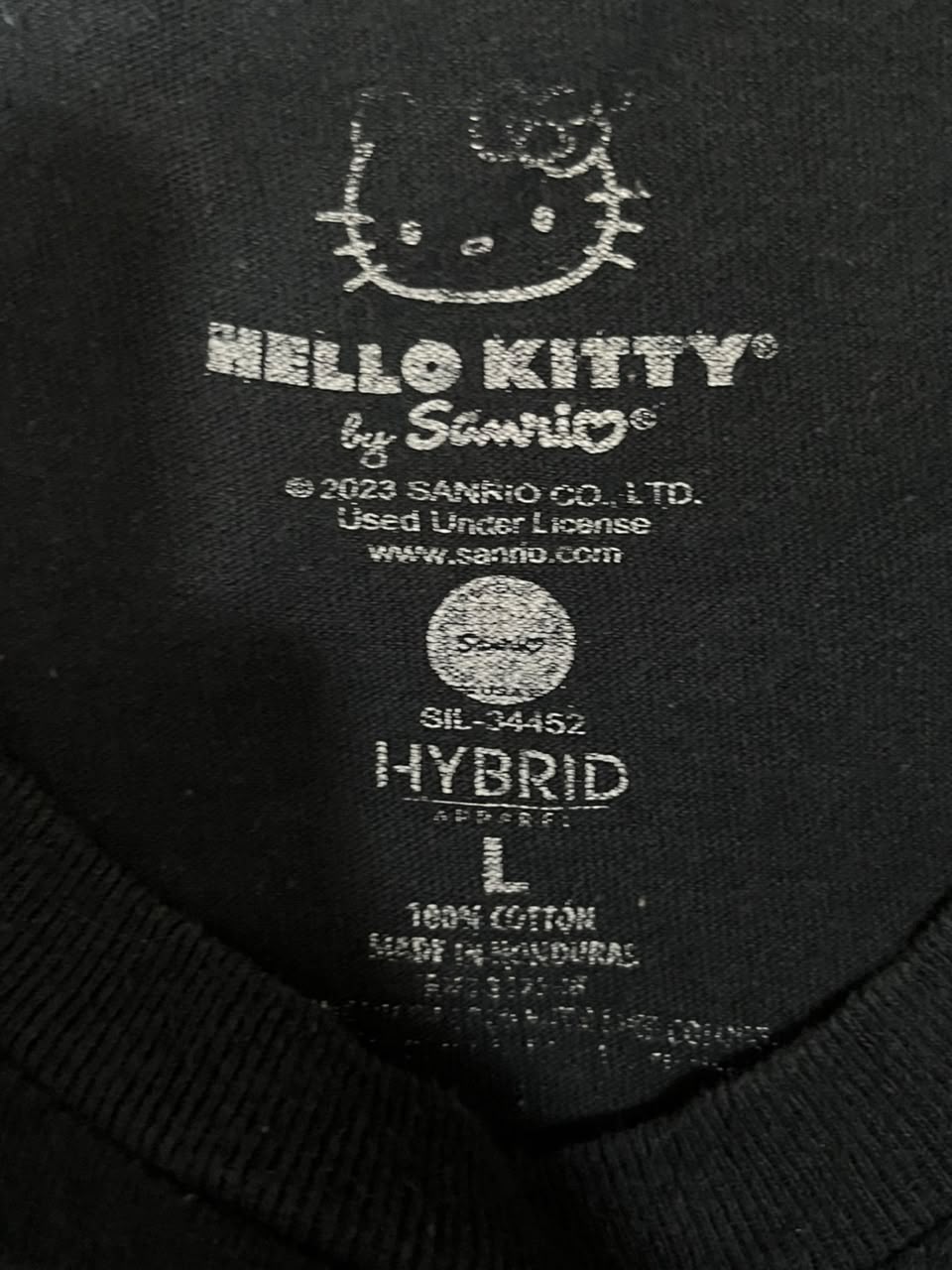 Close-up of the inside neck label of a black Hello Kitty by Sanrio hybrid shirt, showing the Sanrio logo, Hello Kitty face graphic, size L, and 100% cotton details.