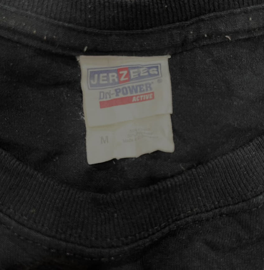 Close-up of a black Jerzees On-Power Active t-shirt neck label, showing the brand logo and size M, with a textured olive green fuzzy surface in the background.