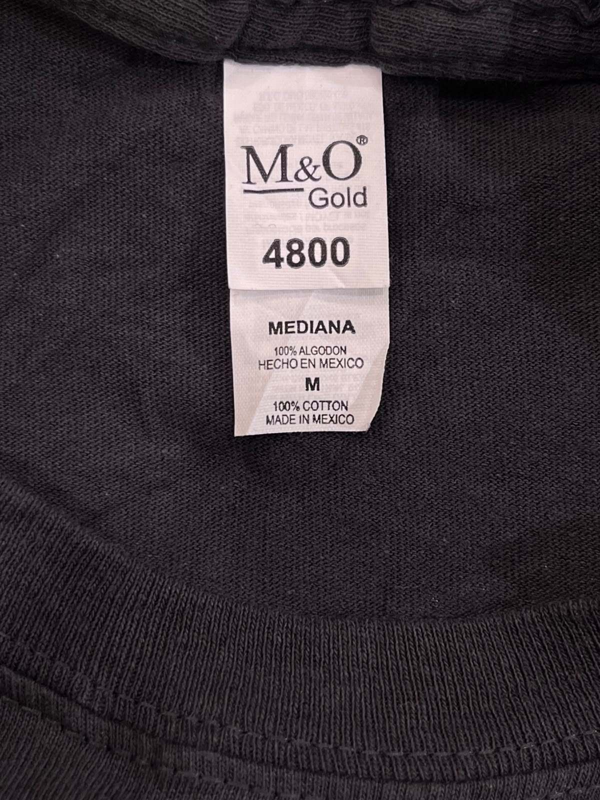 Close-up of a black knit garment neck label reading M&O Gold 4800, size MEDIANA / M, 100% cotton, made in Mexico.