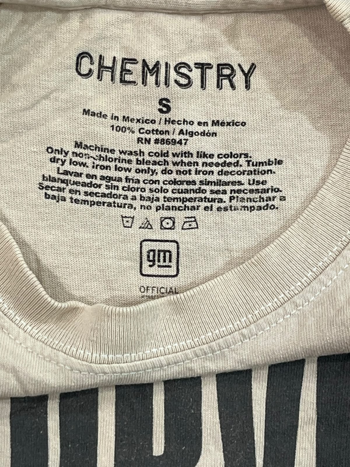Close-up of the inside collar of a Chemistry S crewneck t-shirt, showing the printed inner label with care instructions and a GM Official logo, laid over a black and white striped fabric.