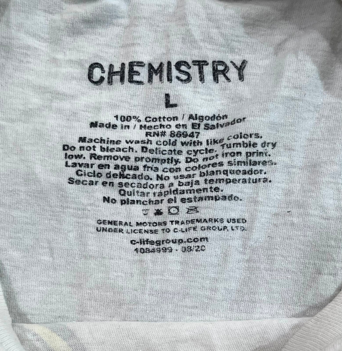 Close-up of a white cotton garment tag with black printed text reading 'CHEMISTRY L' and care instructions, plus C-Life Group branding.
