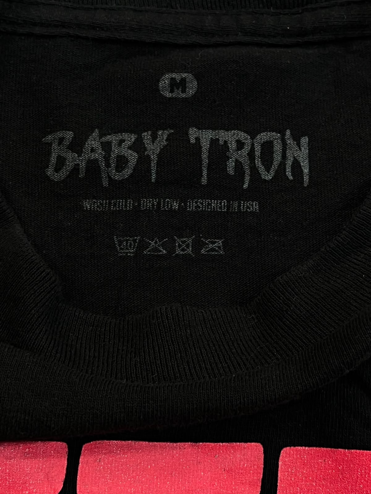 Close-up of the inside neck of a black crewneck t-shirt featuring a gray screen-printed graphic that reads 'BABY TRON' with care instructions below it.