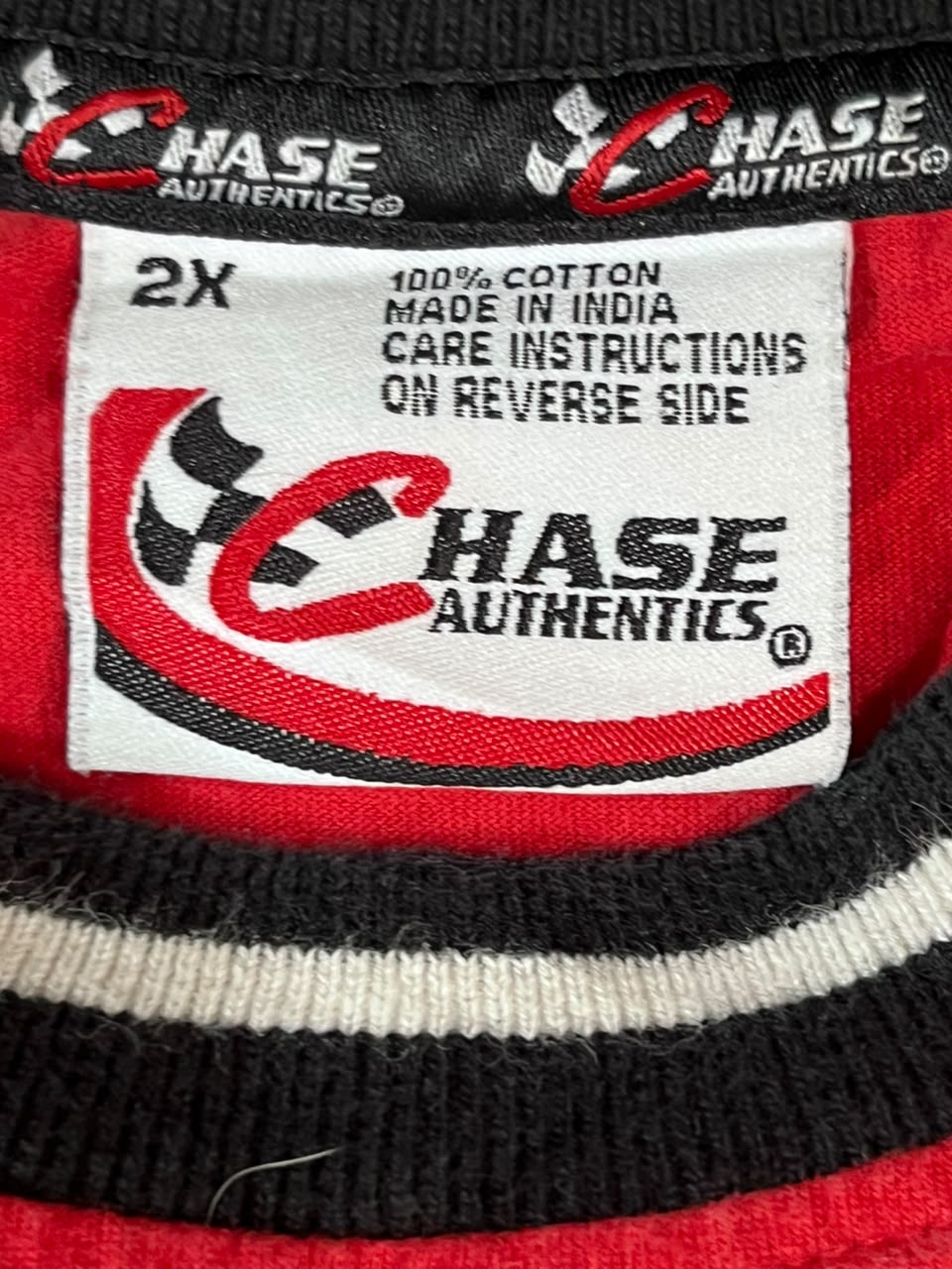 Close-up of a red, black, and white striped knit garment showing the inner neck label with Chase Authentics branding and text reading '2X 100% cotton made in India care instructions on reverse side'.
