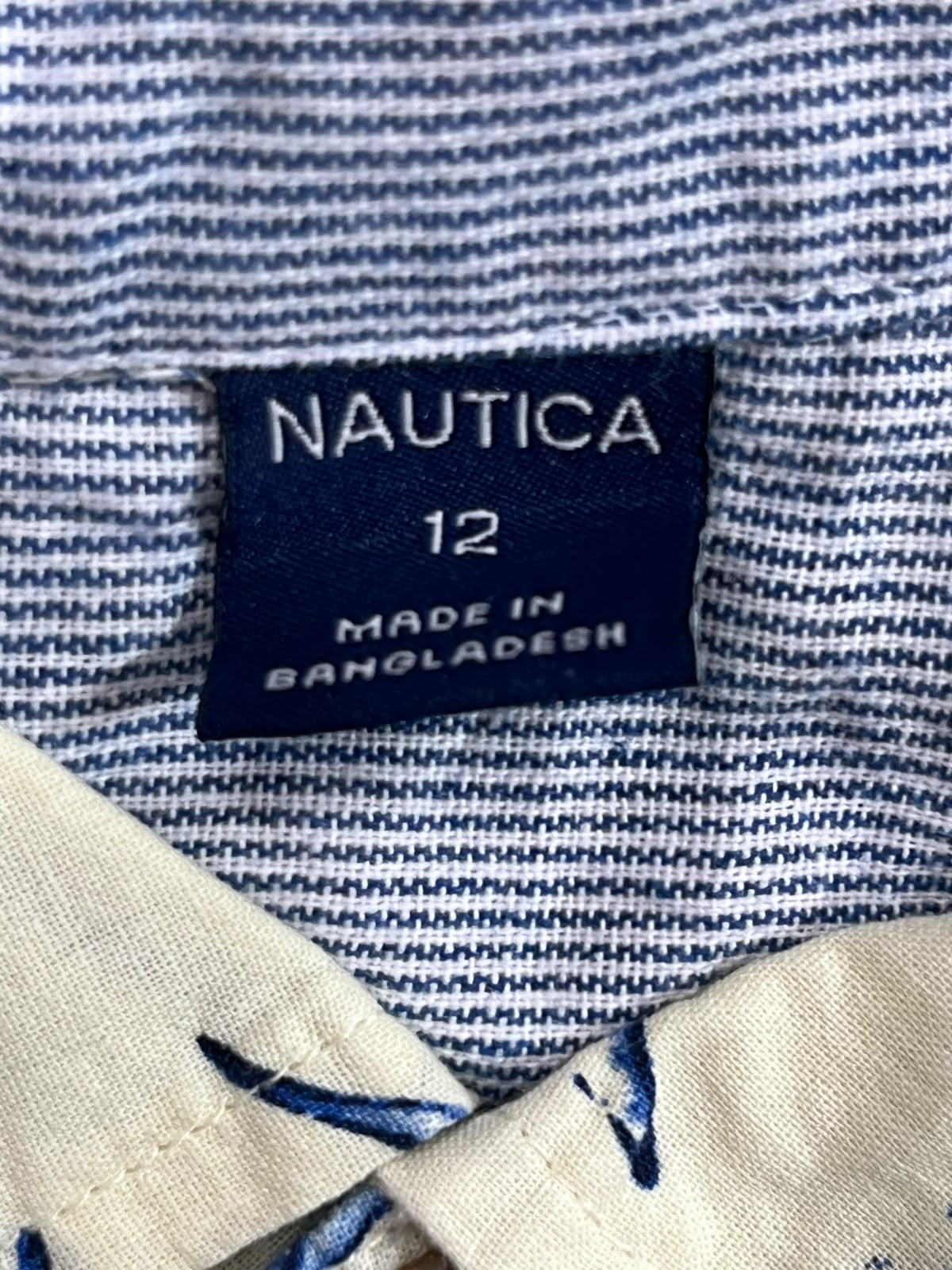 Close-up of a Nautica 12-inch shirt label sewn onto a blue and white striped woven fabric, with a beige collar lining visible at the top.