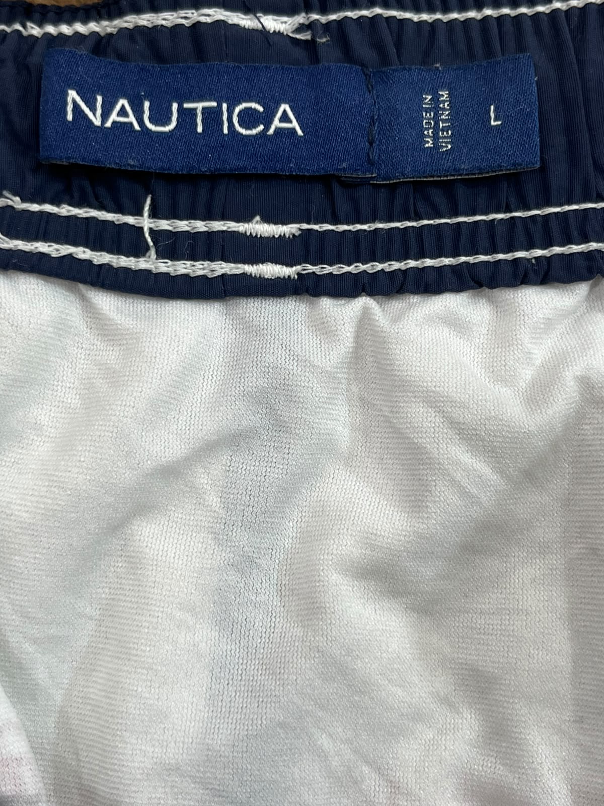 Close-up of a Nautica garment waistband with a navy blue label reading NAUTICA, size L, and white stitching on a white fabric body.