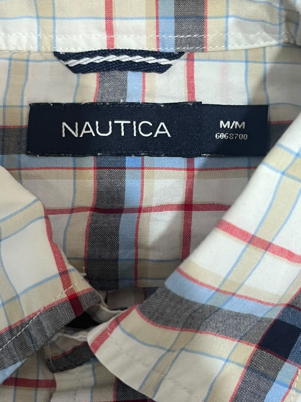 Close-up of a Nautica plaid shirt collar with a black size label reading M/M 6965700. The fabric features a classic plaid pattern in beige, navy, red, and light blue.