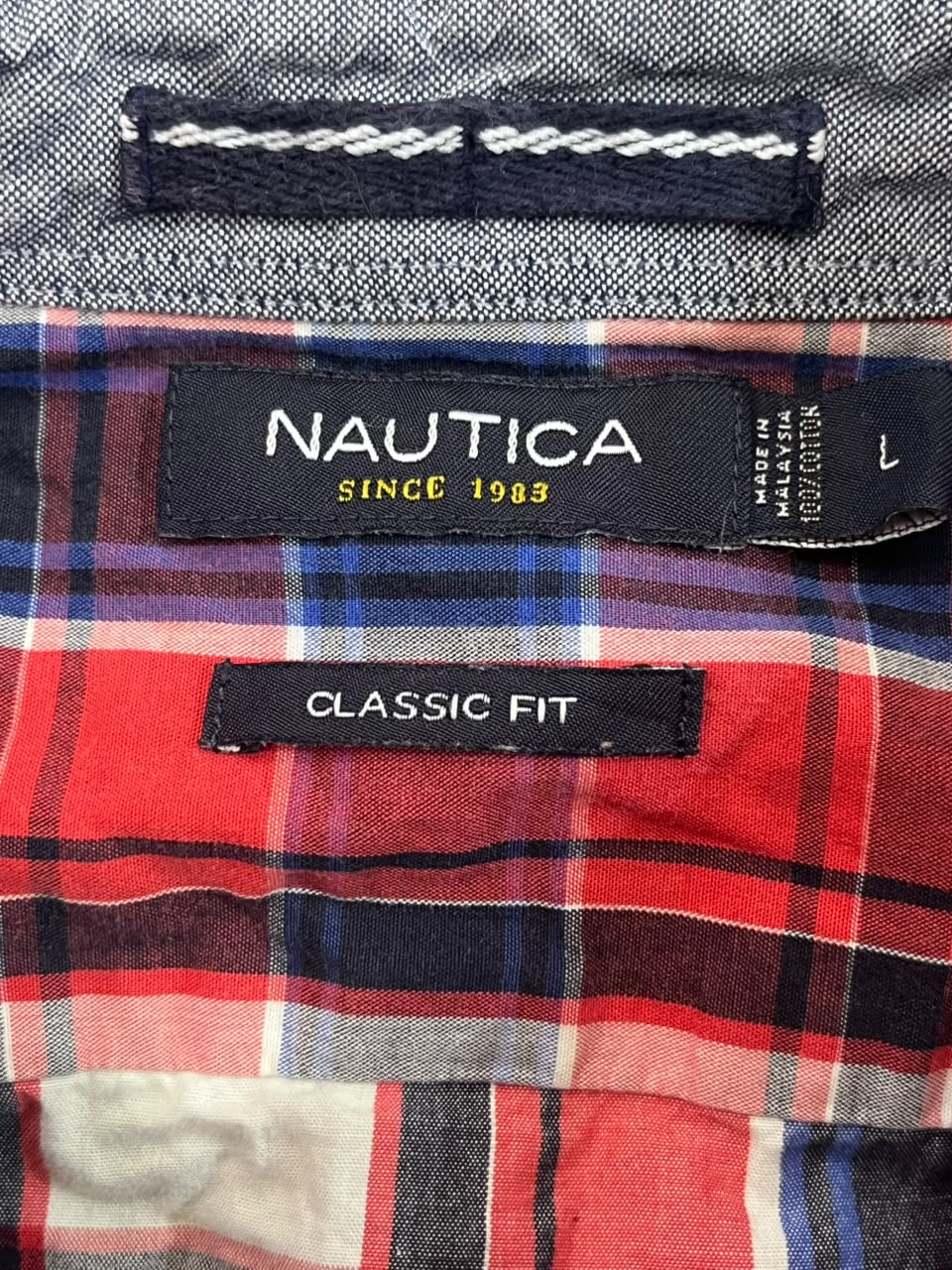 Close-up of a Nautica Classic Fit shirt label on a red, navy, and white plaid garment, with a gray textured fabric section above.