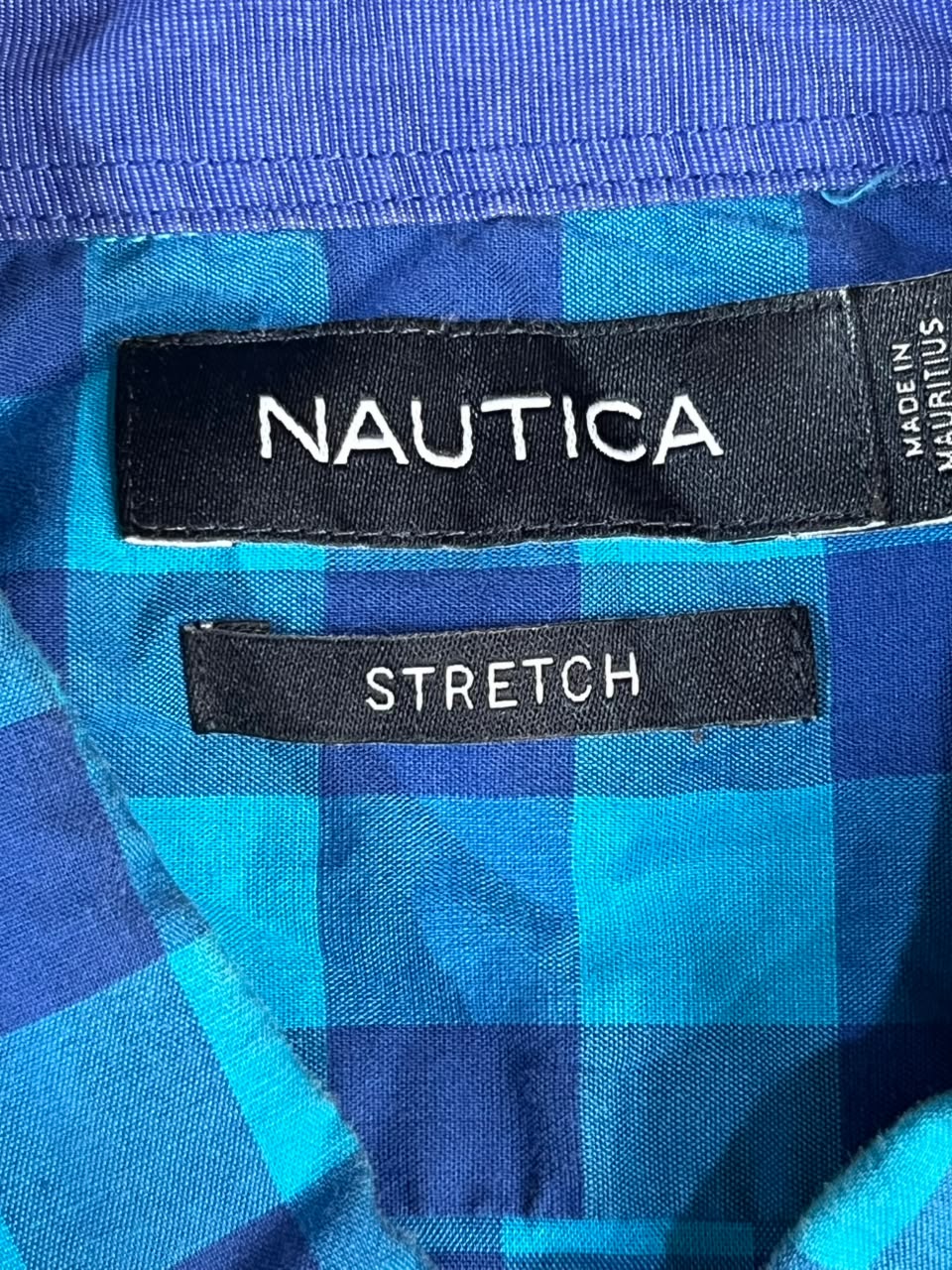 Close-up of a Nautica garment interior showing a blue and turquoise plaid fabric with two black labels: one reading NAUTICA and one reading STRETCH.