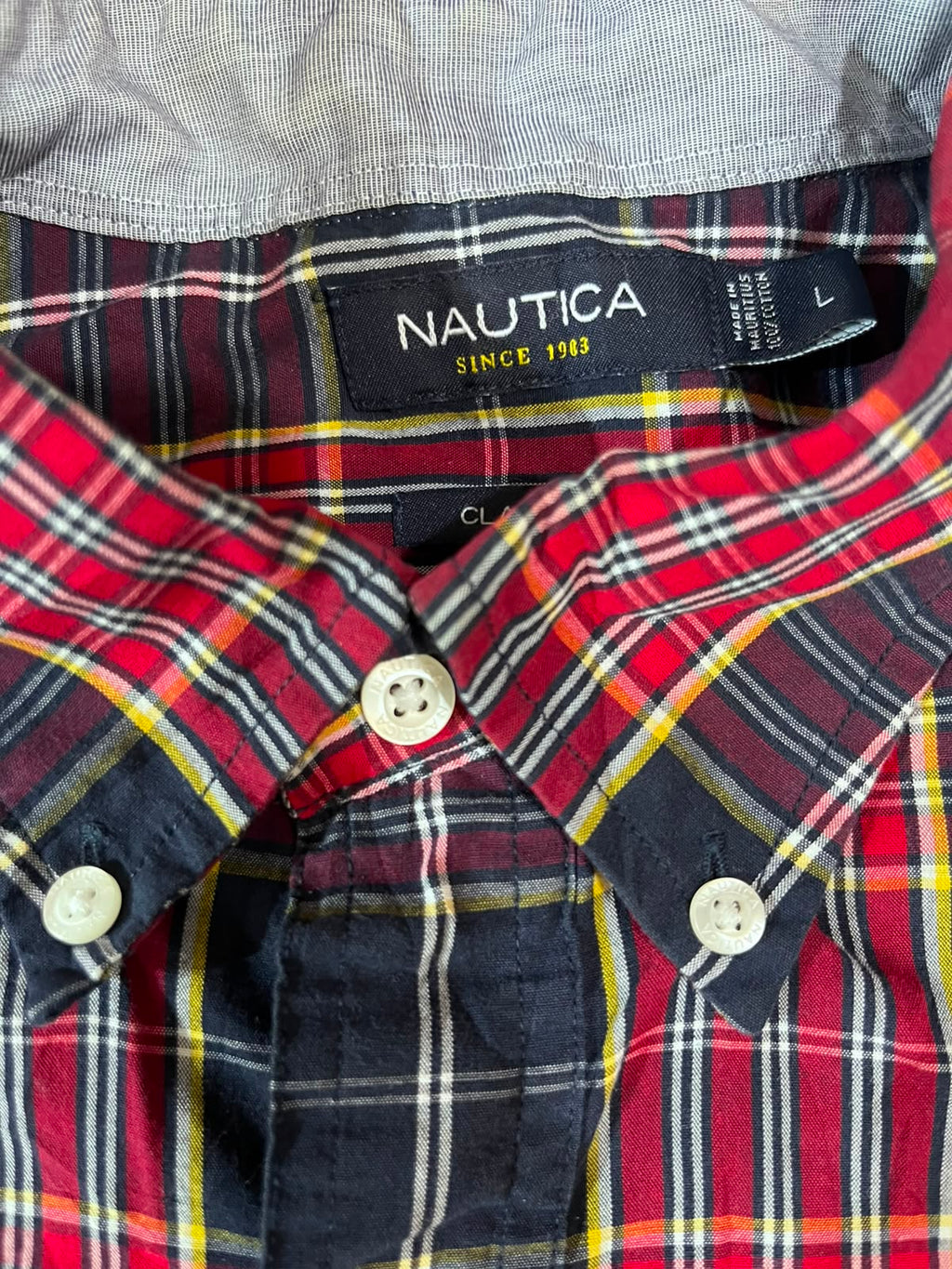 Close-up of a Nautica plaid button-down shirt collar and chest area, showing a red, navy, yellow, and white tartan pattern with a black neck label reading 'NAUTICA SINCE 1993'.