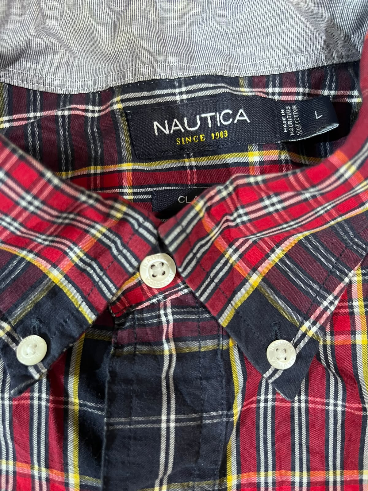 Close-up of a Nautica plaid button-down shirt collar and chest area, showing a red, navy, yellow, and white tartan pattern with a black neck label reading 'NAUTICA SINCE 1993'.