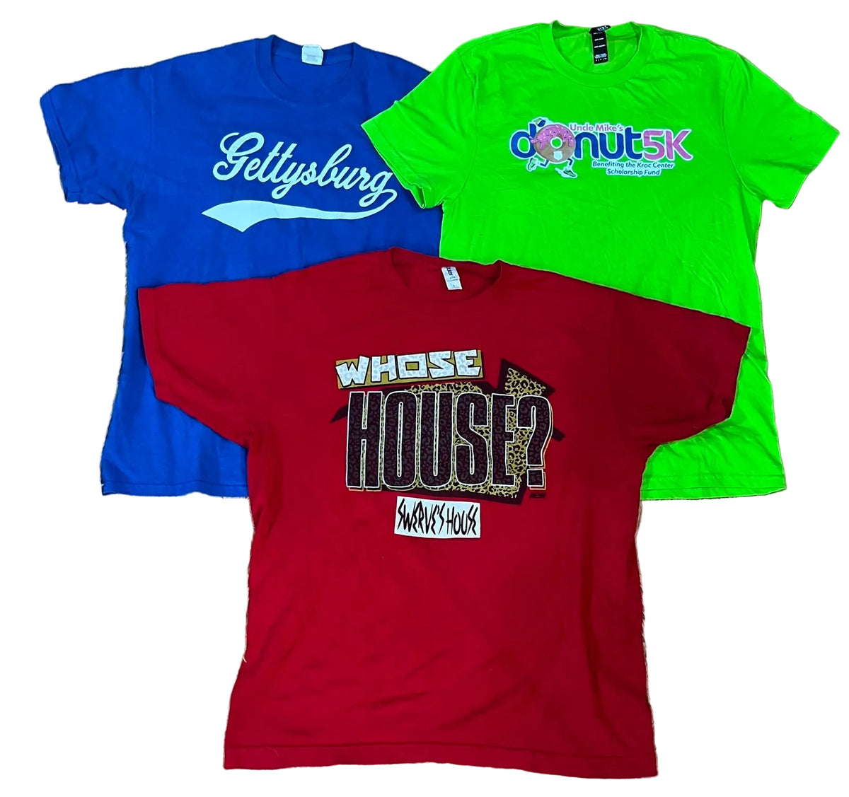 Three vintage graphic t-shirts in blue, green, and red, The blue shirt features 'Sellysburg' in white script, the green shirt has 'donut SK' with a rainbow graphic, and the red shirt displays 'WHOSE HOUSE?' in black and leopard print with a 'Savages House' logo below.