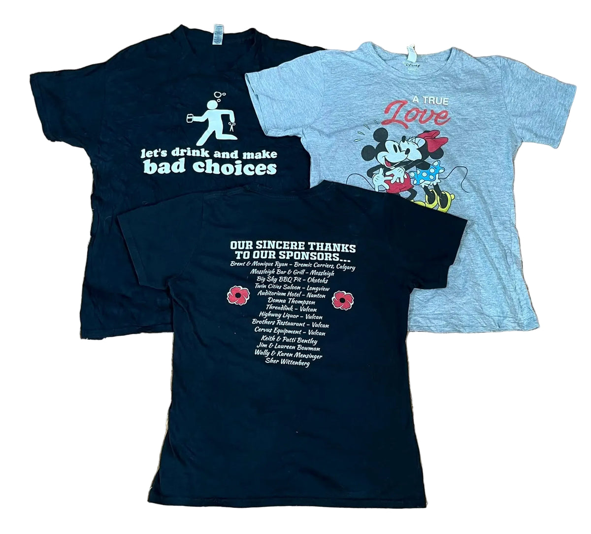 Four graphic t-shirts in black and heather gray. The shirts feature various printed designs including a white running figure on the black shirt, a Mickey and Minnie Mouse graphic with 'A TRUE Love' on the gray shirt, and a sponsor's thank you message on the other black shirt.