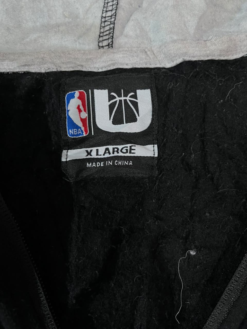 Close-up of the inside neck label of a black garment, showing the NBA logo, a stylized U logo, and the text 'X LARGE MADE IN CHINA'.