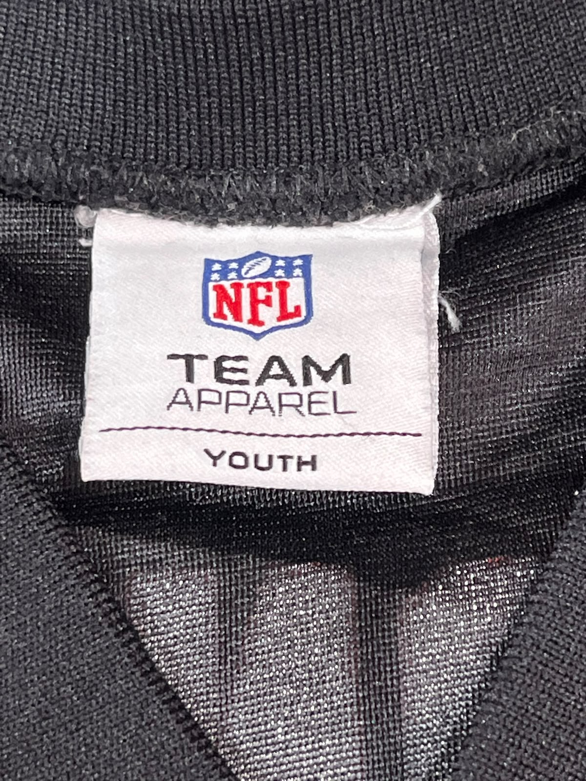 Close-up of a black ribbed knit garment label reading NFL, TEAM APPAREL, and YOUTH.