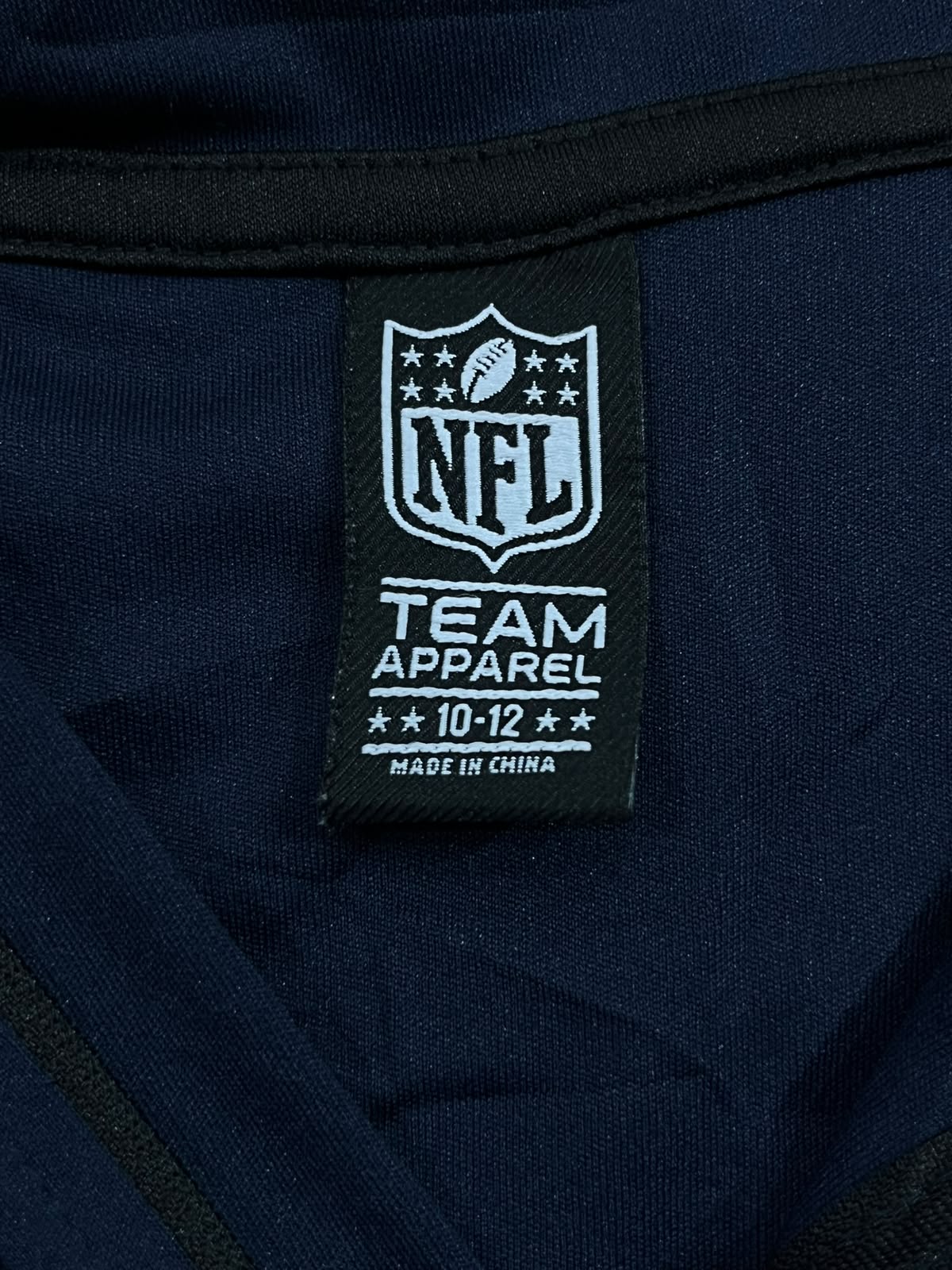 Close-up of a navy blue garment label patch featuring the NFL shield logo and the text 'NFL TEAM APPAREL 10-12 MADE IN CHINA'.